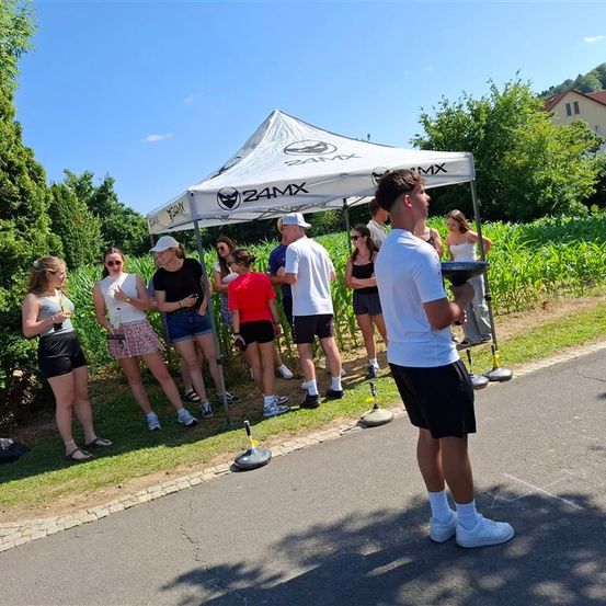 Eine Gruppe von Menschen steht draußen in der Nähe eines Zeltes. Ein Mann in Weiß hält einen Tablett. Die Gruppe trägt verschiedene Farben und steht vor einer Straße.