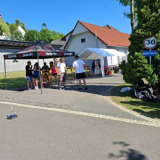 Menschen versammeln sich unter einem Zelt vor einem Haus. Ein Mann steht vor einem Tisch. Ein Schild mit der Nummer 30 ist an einem Pfosten montiert.