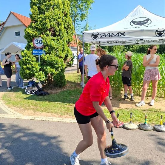 Eine Frau in einem roten Shirt übt den Hammerwurf im Freien in der Nähe eines Zeltes. Im Hintergrund stehen mehrere Personen, einige unter einem Zelt.
