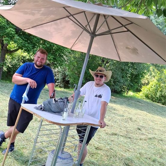 Zwei Männer stehen unter einem großen Schirm. Sie lächeln und scheinen für ein Foto zu posieren. Vor ihnen steht ein Tisch mit Flaschen.