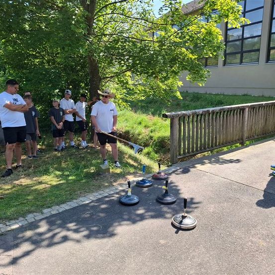 Eine Gruppe von Menschen steht draußen in der Nähe eines Gebäudes und beobachtet einen Mann, der Cornhole spielt. Der Platz hat mehrere Cornhole-Bälle darauf.