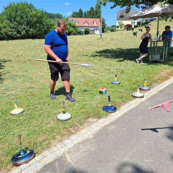 Ein Mann in einem blauen T-Shirt bereitet sich darauf vor, einen Stock bei einem Rasenspiel zu werfen. Eine Frau und ein Mann stehen in der Nähe eines Tisches mit einer Flasche unter einem Schirm.