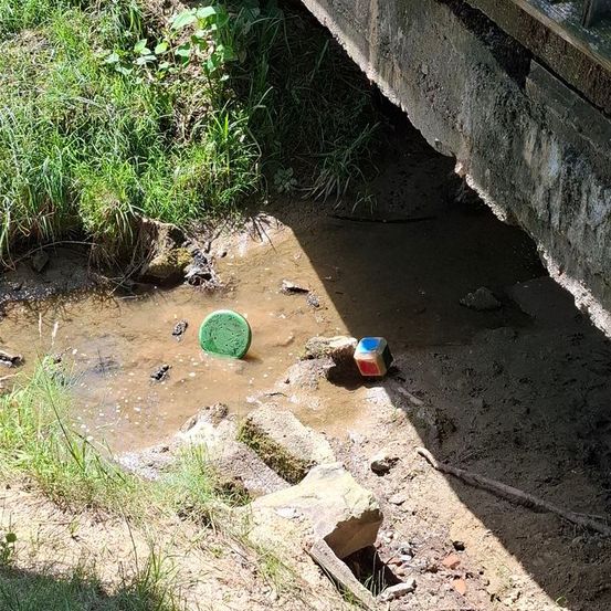 Unter einer Brücke steht stehendes, schlammiges Wasser mit zwei schwimmenden Spielzeugen. Eines ist grün und das andere ist ein bunter Würfel. Es gibt einige Steine und Gras an der Seite.