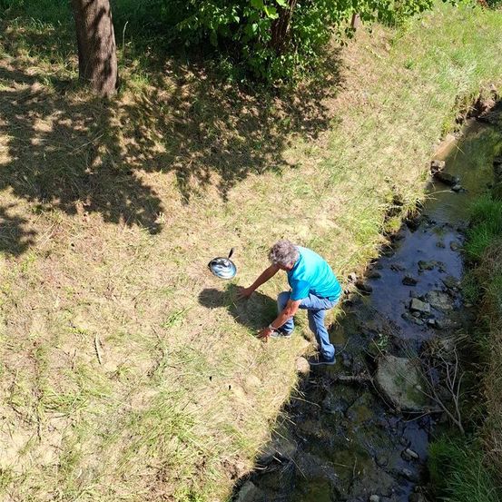 Ein älterer Mann kniet am Ufer eines kleinen Bachs und hält eine Pfanne, möglicherweise zum Fischen. Das Gebiet ist von Gras und Bäumen umgeben.