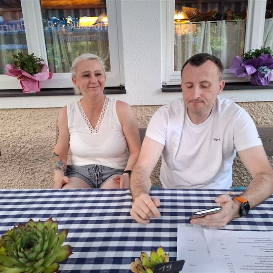 Ein Mann und eine Frau sitzen an einem Tisch mit kariertem Tischtuch. Sie schauen auf ein Handy und ein Papier. Topfpflanzen stehen auf dem Tisch und im Hintergrund.