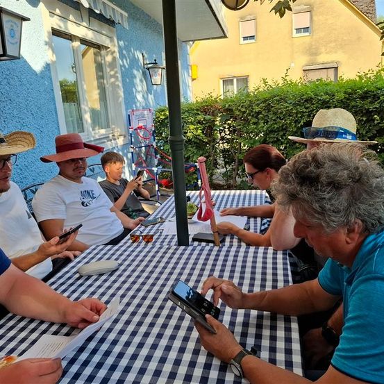 Eine Gruppe von Menschen sitzt um einen Tisch in einem Garten, trägt Hüte und benutzt ihre Handys. Eine Frau liest, und es gibt ein Gebäude, Büsche und einen Basketballkorb im Hintergrund.