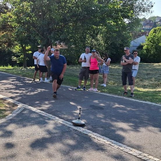 Eine Gruppe von Menschen versammelt sich auf einem Weg in einem Park und beobachtet einen Mann in einem blauen T-Shirt, der einen Frisbee wirft. Einige Leute stehen auf dem Gras, während andere auf dem Bürgersteig stehen.