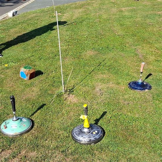 Das Bild zeigt ein Grasfeld mit vier Objekten: einem kleinen Würfel, drei Rollen und einem Pfahl. Der Würfel hat blaue und grüne Seiten. Die Rollen sind grün, blau und gelb.