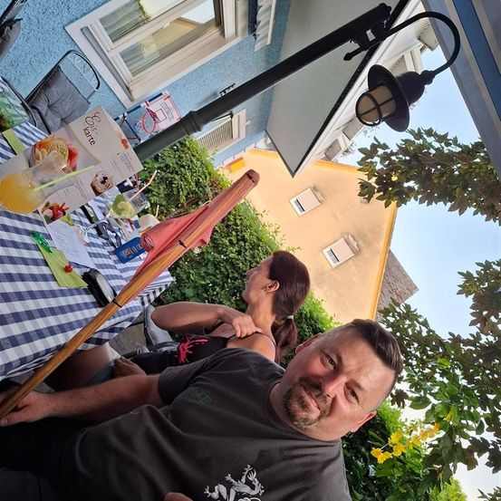 Zwei Personen sitzen auf einer Terrasse unter einer Lampe. Ein Mann lächelt für die Kamera, während eine Frau hinter ihm ruht. Ein Tisch hält Getränke, und eine Box 'Espresso Karte' steht in der Nähe. Gebäude mit Fenstern und Pflanzen sind im Hintergrund.