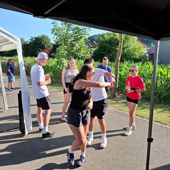 Eine Gruppe von Menschen steht auf einem Weg mit einem Baldachin darüber. Sie sind in der Nähe eines Maisfeldes. Eine Frau mit Sonnenbrille scheint zu tanzen.
