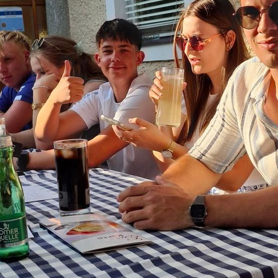 Eine Gruppe von Menschen sitzt an einem Tisch, ein Mann gibt einen Daumen hoch und eine Frau hält ein Glas mit Getränk.