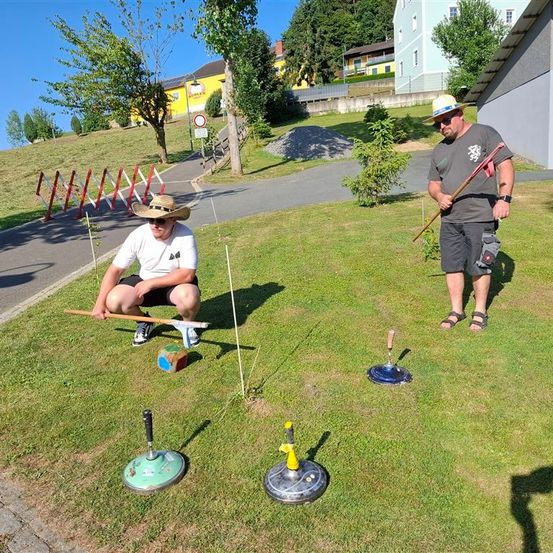 Zwei Männer in Hüten und Sonnenbrillen bereiten sich auf ein Spiel auf einem Rasenfeld vor, halten Stöcke und sind von Spielgeräten umgeben. Hinter ihnen führt eine Straße zu einem gelben Gebäude.