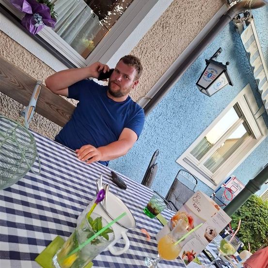 Ein Mann in einem blauen T-Shirt sitzt auf einer Bank vor einem Haus und hält ein Handy am Ohr. Vor ihm steht ein Tisch mit Getränken und einer Speisekarte.