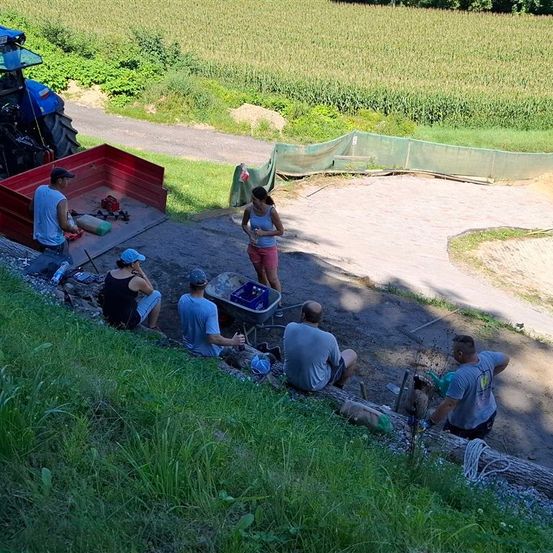 Eine Gruppe von Menschen arbeitet im Freien auf einem Feld. Eine Frau steht mit einem Schubkarren. In der Nähe ist ein Traktor geparkt. Zwei Personen sitzen an einem Hang, andere sitzen auf dem Boden.