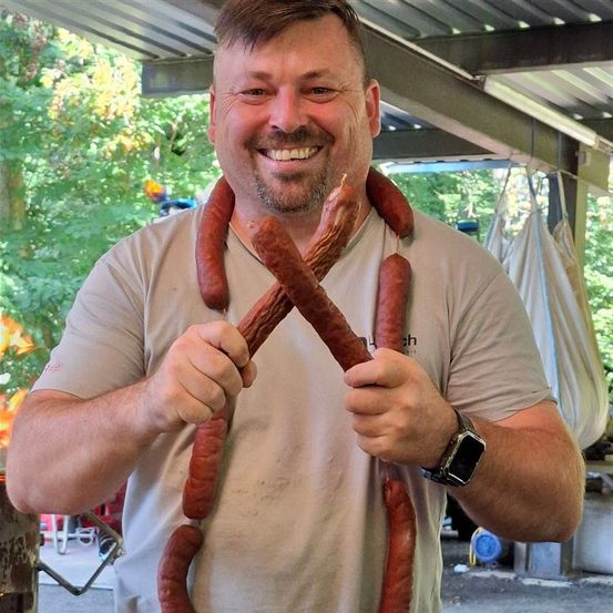 Ein Mann lächelt und hält mehrere Würste um seinen Hals, steht in einem schattigen Außenbereich mit Bäumen im Hintergrund.