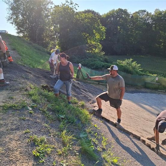 Eine Gruppe von Menschen arbeitet an einem Schotterweg mit einer Steigung, trägt Handschuhe und Mützen. Der Weg ist von Gras und Bäumen umgeben.