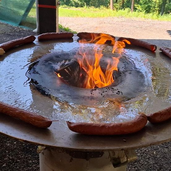 Ein Outdoor-Kochplatz mit einem runden Grill, auf dem Würste um einen zentralen Feuerkorb herum angeordnet sind, auf einem Kiesbereich mit Grün im Hintergrund.