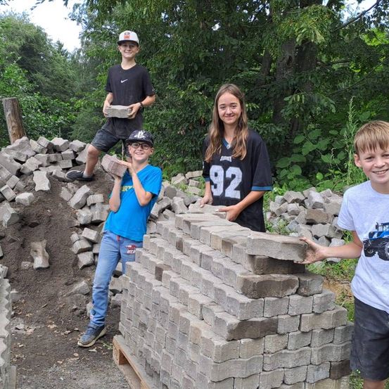 Vier Kinder posieren mit Ziegeln und einem Stapel, lächeln in einem sonnigen, bewaldeten Bereich. Bäume und Grünpflanzen umgeben sie.
