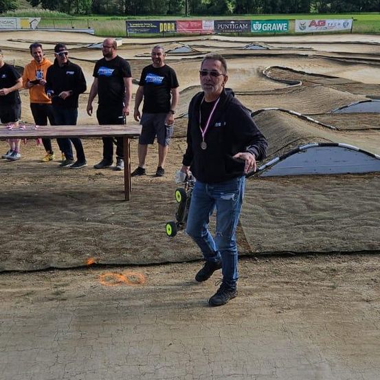 Ein Mann in einem schwarzen Jacken hält ein ferngesteuertes Auto und steht in einem staubigen Bereich, während andere zusehen. Vor ihnen steht ein Holztisch.