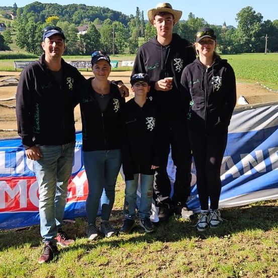 Fünf Personen in passenden schwarzen Jacken posieren für ein Foto auf einem Grasfeld mit einem blauen Banner hinter ihnen. Bäume und Hügel sind im Hintergrund.