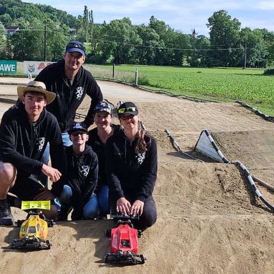 Fünf Personen posieren für ein Foto auf einer Erdstrecke. Sie tragen alle schwarze Kapuzenpullover. Sie halten zwei ferngesteuerte Autos. Ein Schild ist an einem Pfosten montiert. Im Hintergrund befindet sich ein Zaun. Auf der linken Seite ist ein Zaun.