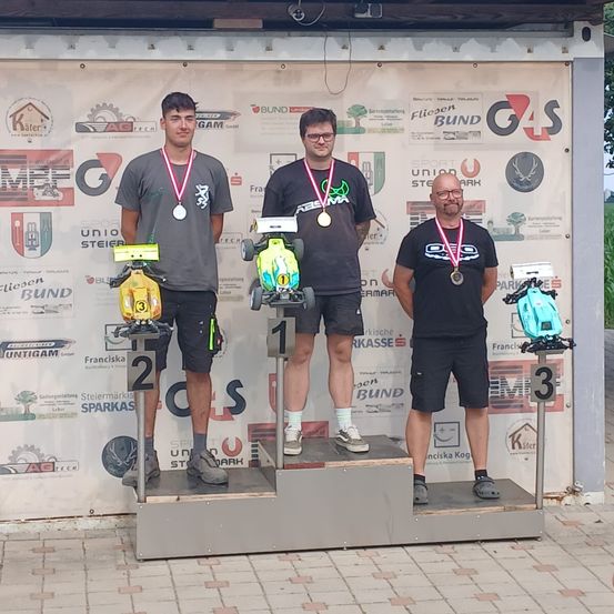 Drei Männer stehen auf einem Podest, alle tragen Medaillen. Der linke Mann hält ein kleines Automodell, der mittlere Mann hält ein Spielzeugauto und der rechte Mann hält einen Spielzeughubschrauber. Hinter ihnen ist eine weiße Wand mit verschiedenen Logos und Texten.