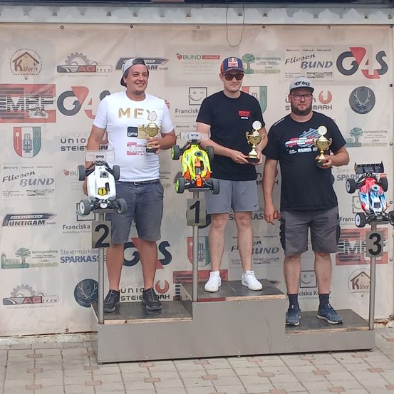 Drei Männer stehen auf einem Podest, jeder hält eine Trophäe. Sie lächeln und scheinen für ein Foto zu posieren. Der Hintergrund besteht aus Bannern mit Logos und Text.