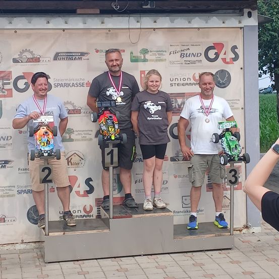 Vier Personen stehen mit Medaillen auf einem Podest, zwei Männer an den Seiten und zwei in der Mitte, eine Frau und ein Mann. Sie alle lächeln und halten Spielzeugautos in den Händen.