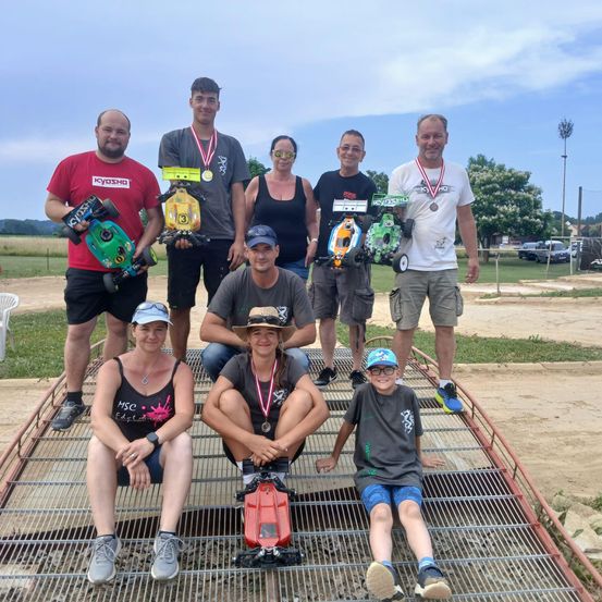 Eine Gruppe von Menschen posiert mit ihren Spielzeugautos und Medaillen für ein Foto. Sie stehen auf einer Metallplattform und sind von einem Feld mit Erde umgeben.