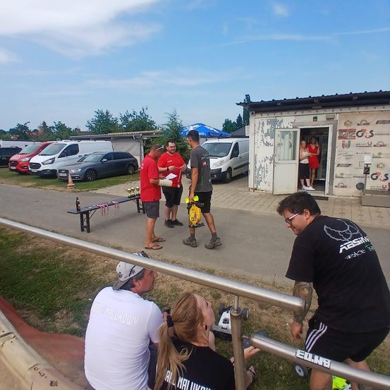 Eine Gruppe von Menschen steht neben einem kleinen Gebäude, in dem zwei Frauen sind. Ein Mann hält ein Papier und ein anderer einen gelben Beutel. Einige Autos sind geparkt.