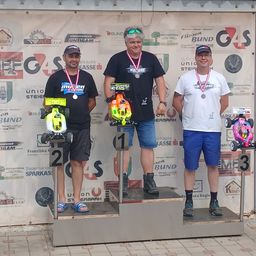 Drei Männer stehen auf einem Podest mit Medaillen um den Hals und halten ferngesteuerte Autos. Sie tragen lässige Sommerkleidung. Dahinter ist eine Kulisse mit Logos und Text.