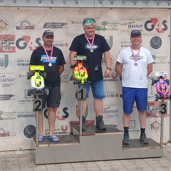 Drei Männer stehen auf einem Podest mit Medaillen um den Hals und halten ferngesteuerte Autos. Sie tragen lässige Sommerkleidung. Dahinter ist eine Kulisse mit Logos und Text.