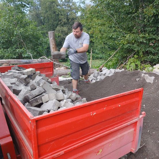 Ein Mann in einem grauen T-Shirt und Shorts benutzt einen Spaten, um einen roten Container mit Steinen und Erde zu beladen.