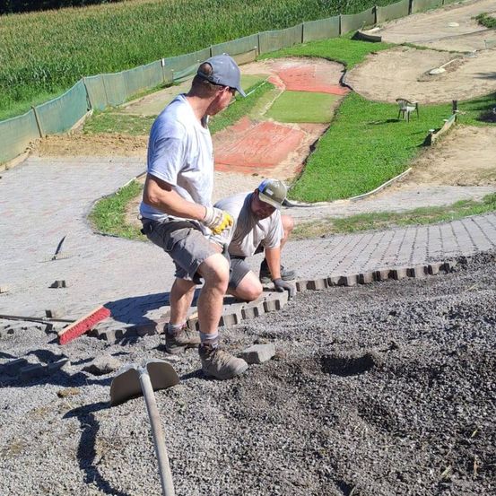 Zwei Männer arbeiten an einem Kiesweg, einer mit einer Schaufel, der andere kniet. Sie sind von grünem Gras und einem Zaun umgeben.