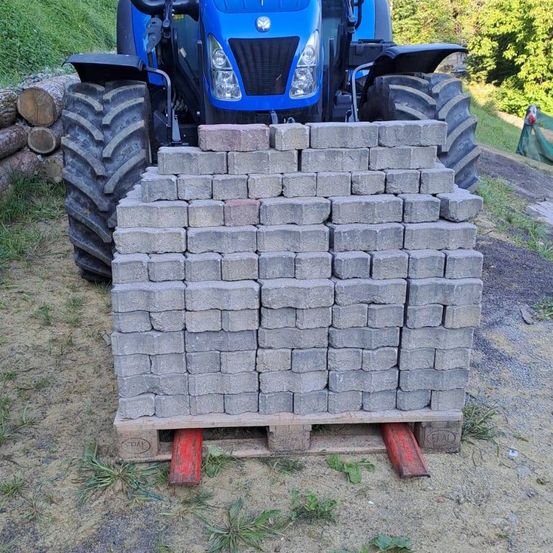 Ein blauer Traktor steht hinter einem großen Stapel grauer Betonblöcke auf einem Holzpalette im Freien.
