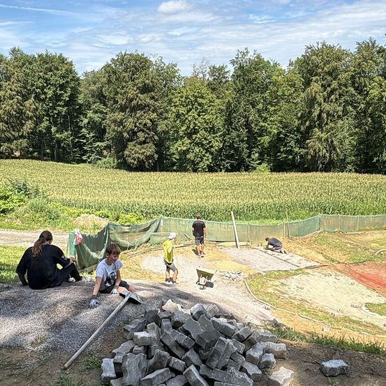 Eine Gruppe von Menschen arbeitet an einem Projekt in einer ländlichen Gegend. Einige sitzen auf dem Boden, während andere stehen. Vor ihnen liegen Steinhaufen. Im Hintergrund steht ein Netzzaun. Dahinter befindet sich ein weitläufiges Feld und Bäume.