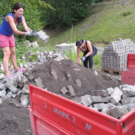 Zwei Frauen sortieren Schutt und Trümmer, wahrscheinlich Teil eines Abrissprojekts. Eine Frau hält eine Schaufel, während die andere Handschuhe trägt und Steine aufhebt. Ein roter LKW befindet sich in der Nähe, möglicherweise zum Transport des Schutts.