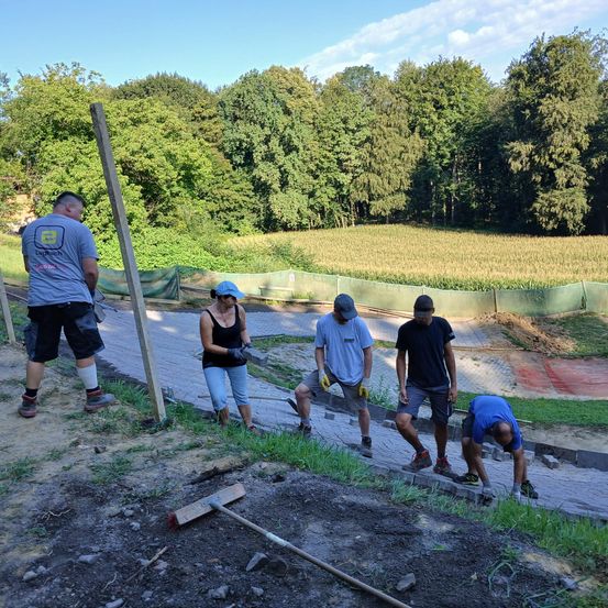 Fünf Arbeiter legen Ziegel auf einer Baustelle, mit einem Wald und Feld im Hintergrund. Ein Arbeiter hält eine Kelle, und ein anderer beugt sich.