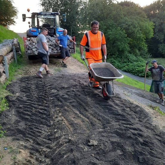 Vier Männer arbeiten auf einem Pfad, im Hintergrund steht ein Traktor. Ein Mann schiebt eine Schubkarre mit Kies.