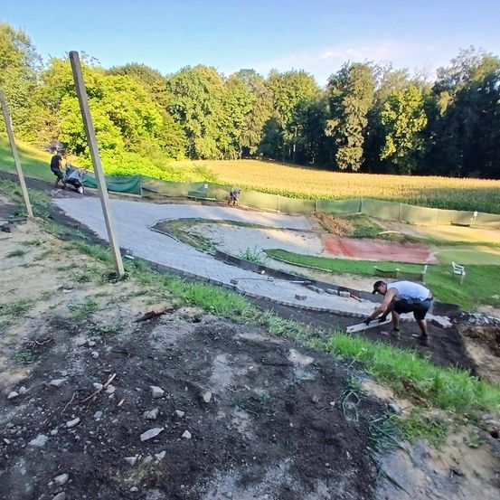 Mehrere Arbeiter bauen einen kurvigen Pfad auf einem Hügel, mit einem großen Feld und Bäumen im Hintergrund. Ein Arbeiter ist auf einem Roller, während andere am Pfad arbeiten.