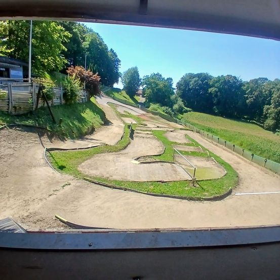 Blick von innen aus einem Fahrzeug auf eine kurvenreiche Schotterpiste mit grünem Gras, Kurven und einem entfernten Haus mit Holzzaun unter einem klaren Himmel.