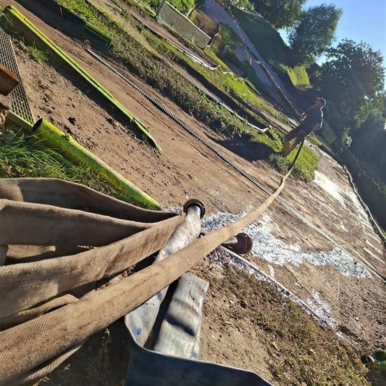 Ein Mann arbeitet im Freien an einem sonnigen Tag und hält einen Schlauch, der Wasser sprüht. Verschiedene Werkzeuge und Rohre sind auf dem mit Erde bedeckten Boden verstreut. Bäume und Gras sind im Hintergrund zu sehen.