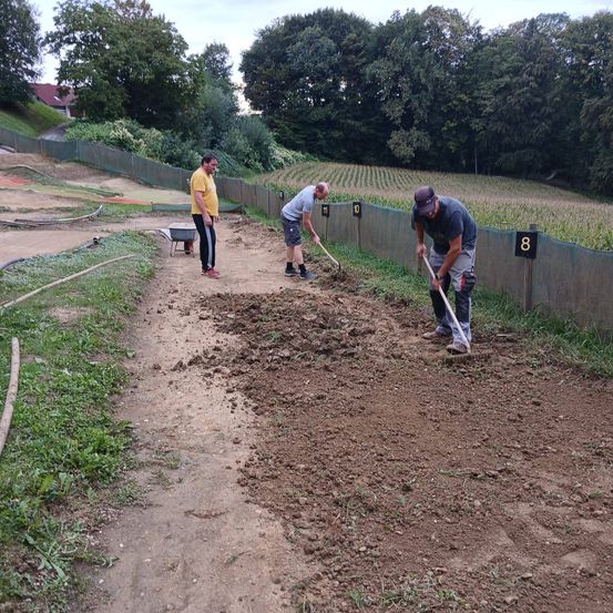 Drei Männer arbeiten auf einem schlammigen Gelände, möglicherweise zur Vorbereitung eines Feldes. Einer steht in der Nähe eines Schubkarrens, die anderen beiden benutzen Schaufeln. Nummerierte Schilder befinden sich an der Wand.