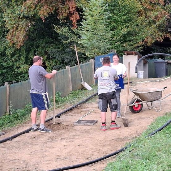 Drei Personen arbeiten in einem Gartenbereich. Zwei Männer halten Schaufeln, während eine Frau in der Nähe steht. Sie sind von Werkzeugen und Schubkarren umgeben.