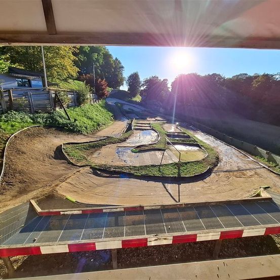 Eine Dirtbike-Strecke wird von oben mit strahlend heller Sonne gesehen. Sie hat Kurven, Wendungen und einen geraden Weg. Links eine Holzumzäunung und etwas Grün. Die Strecke ist nass, was auf kürzlichen Regen hinweist.