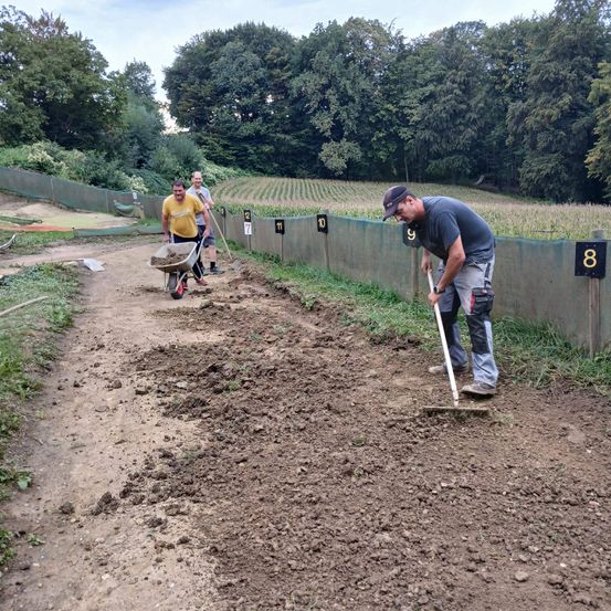 Männer arbeiten auf einem Erdweg, einer schiebt einen Schubkarren und der andere benutzt eine Harke. Die Zahlen 8, 9, 10, 11 und 12 sind an einer Wand markiert, und ein Feld mit Pflanzen ist im Hintergrund zu sehen.