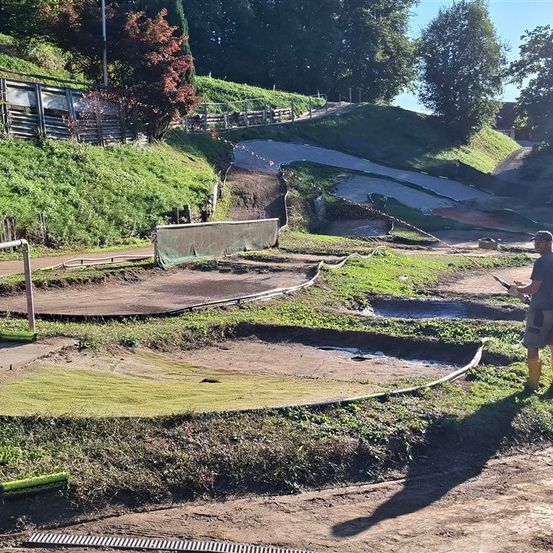 Ein Mann steht und hält ein Werkzeug auf einer Erdoberfläche. Er trägt einen Hut und Stiefel. Das Gebiet ist von einem Zaun umgeben, und es gibt Bäume und Pflanzen. Rechts befindet sich ein Wassersprüher. In der Ferne gibt es eine Rampe mit einer Steigung.