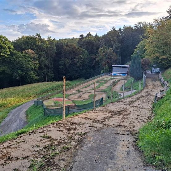 Ein mit grünem Gras umgebener Erdweg führt zu einer Motocross-Strecke. Im Hintergrund ein Gebäude mit weißem Dach und einem grünen Feld.