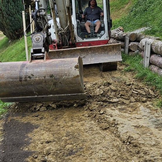Ein Mann bedient einen rot-weißen Bagger, der Erde und Steine bewegt. Er sitzt im Fahrersitz, trägt eine Halskette und Turnschuhe.
