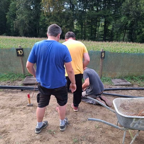 Drei Männer arbeiten auf einer Baustelle mit den Nummern 9 und 10. Einer kniet und hält ein Werkzeug, während die anderen beobachten. Ein Schubkarren und ein Feld sind in der Nähe.